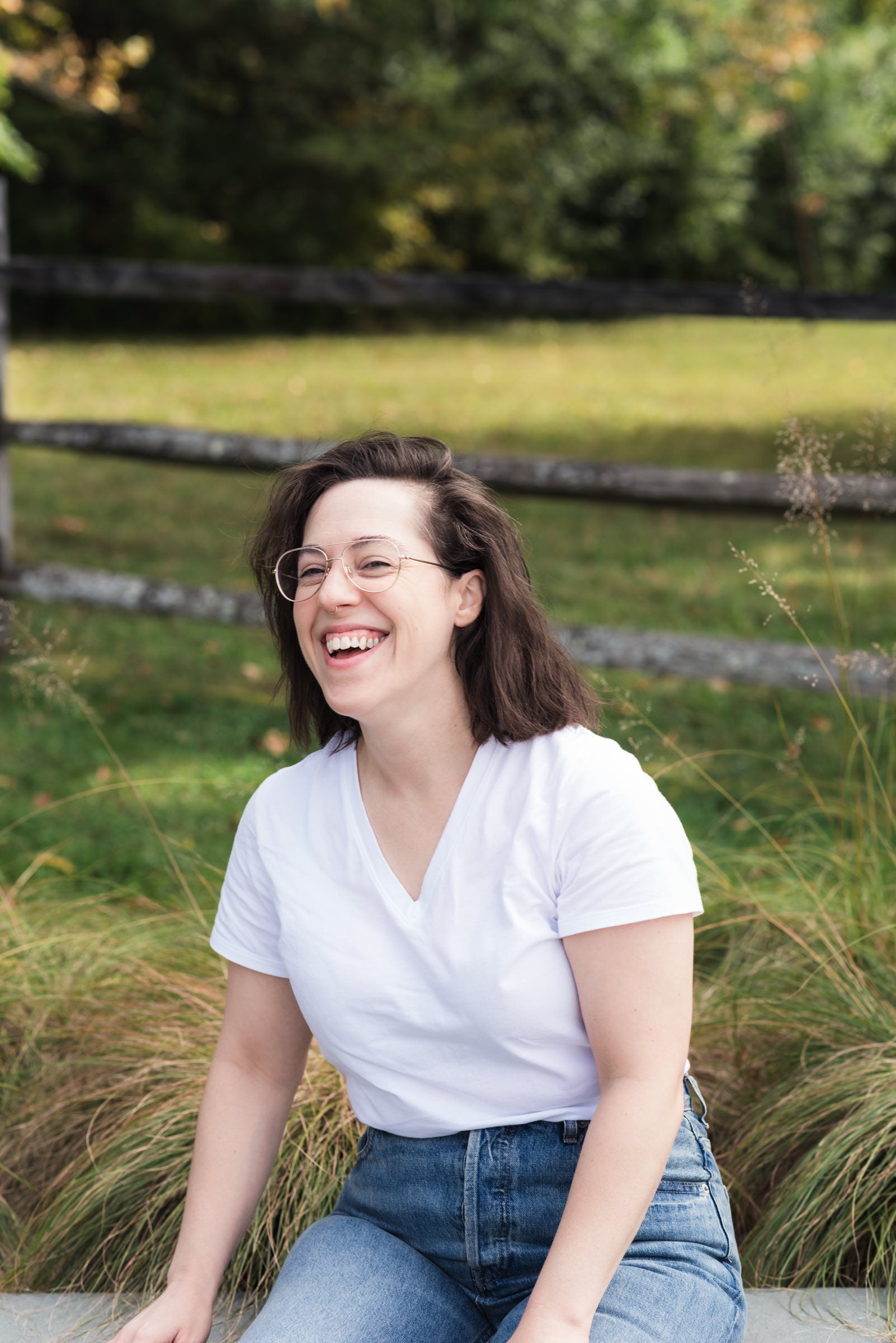 A woman with brown hair and glasses, wearing a white V-neck t-shirt and blue jeans, sitting outdoors in a grassy field with a wooden fence in the background.