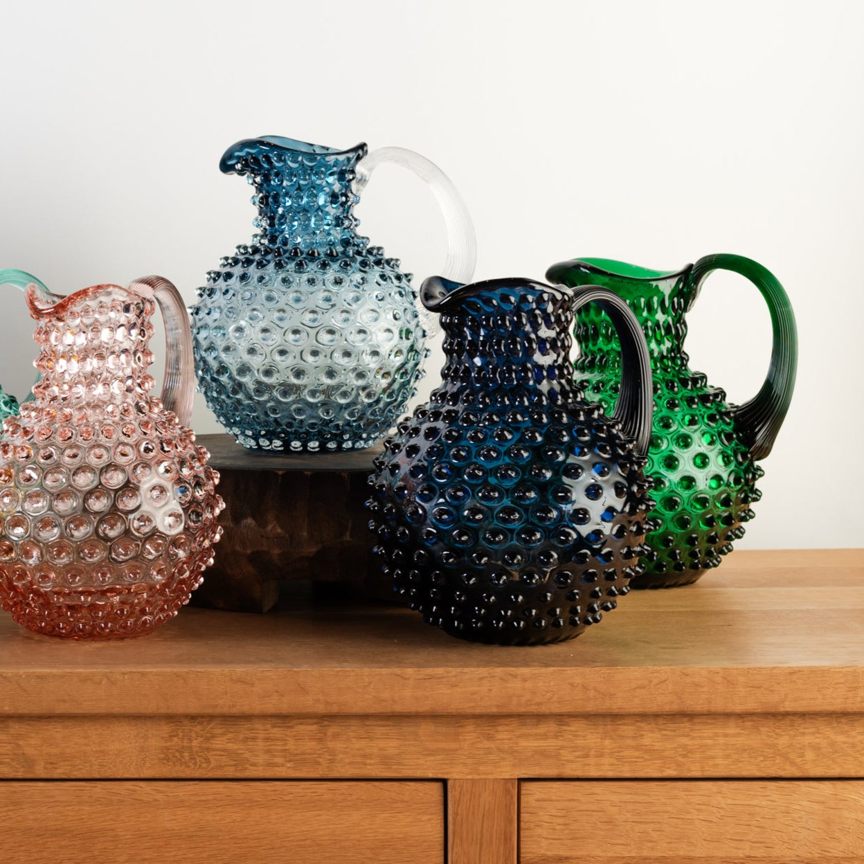 Collection of glass hobnail pitchers on a wood table in front of a white wall at Addison West