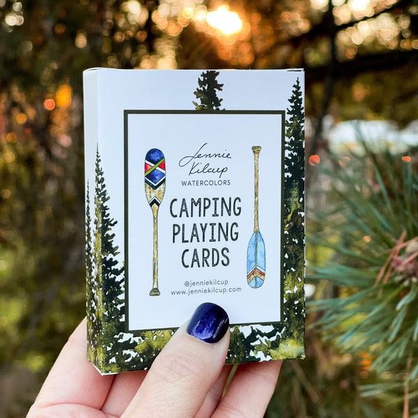 Pack of camping playing cards with watercolor painted images held by a hand in front of trees