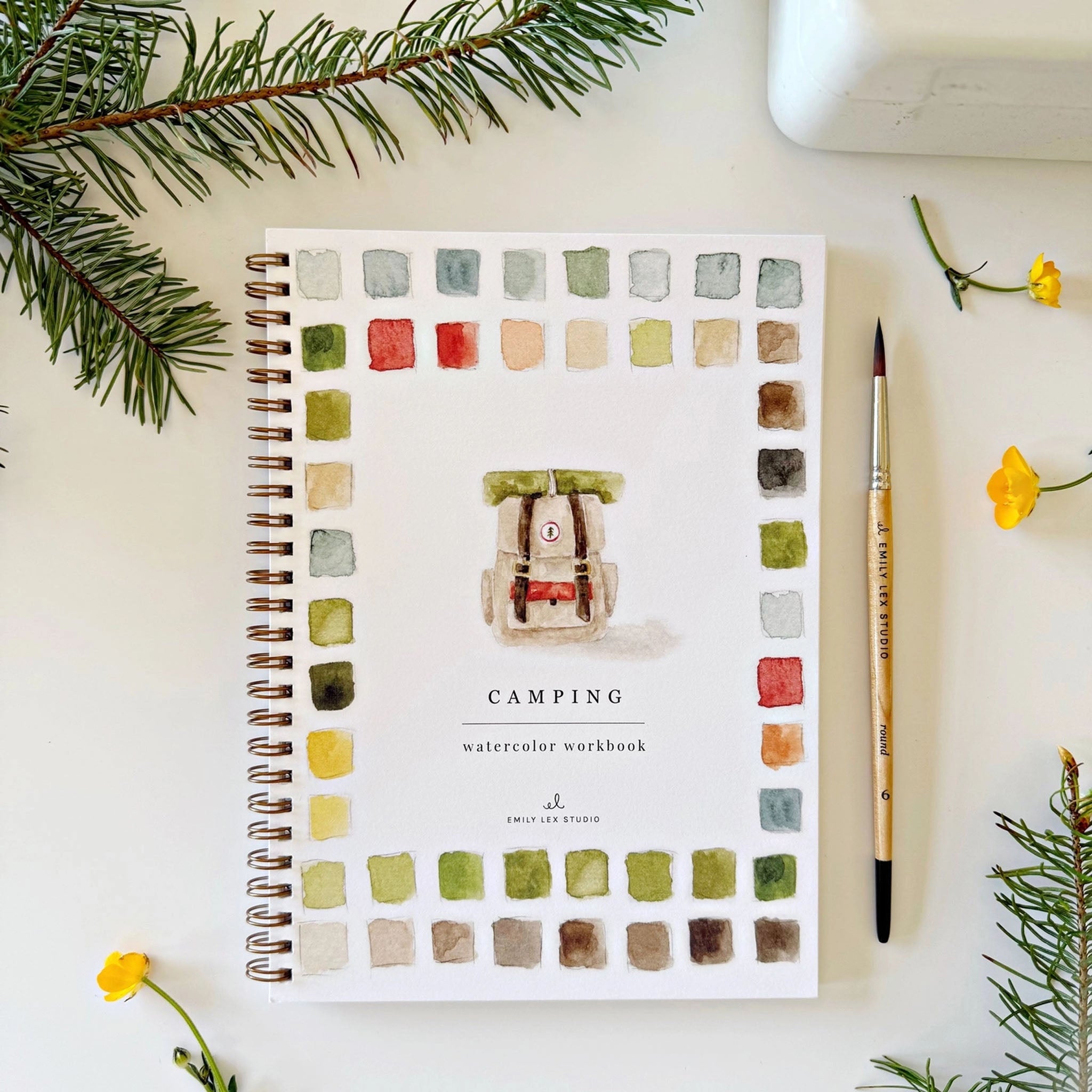 Emily Lex Camping Watercolor Workbook on a table with flowers and a paint brush