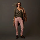 A woman stands against a dark gray studio background wearing a dark olive green V-neck sweater, dusty pink pants, dark brown loafers, and a leopard-print coat draped over her shoulder.
