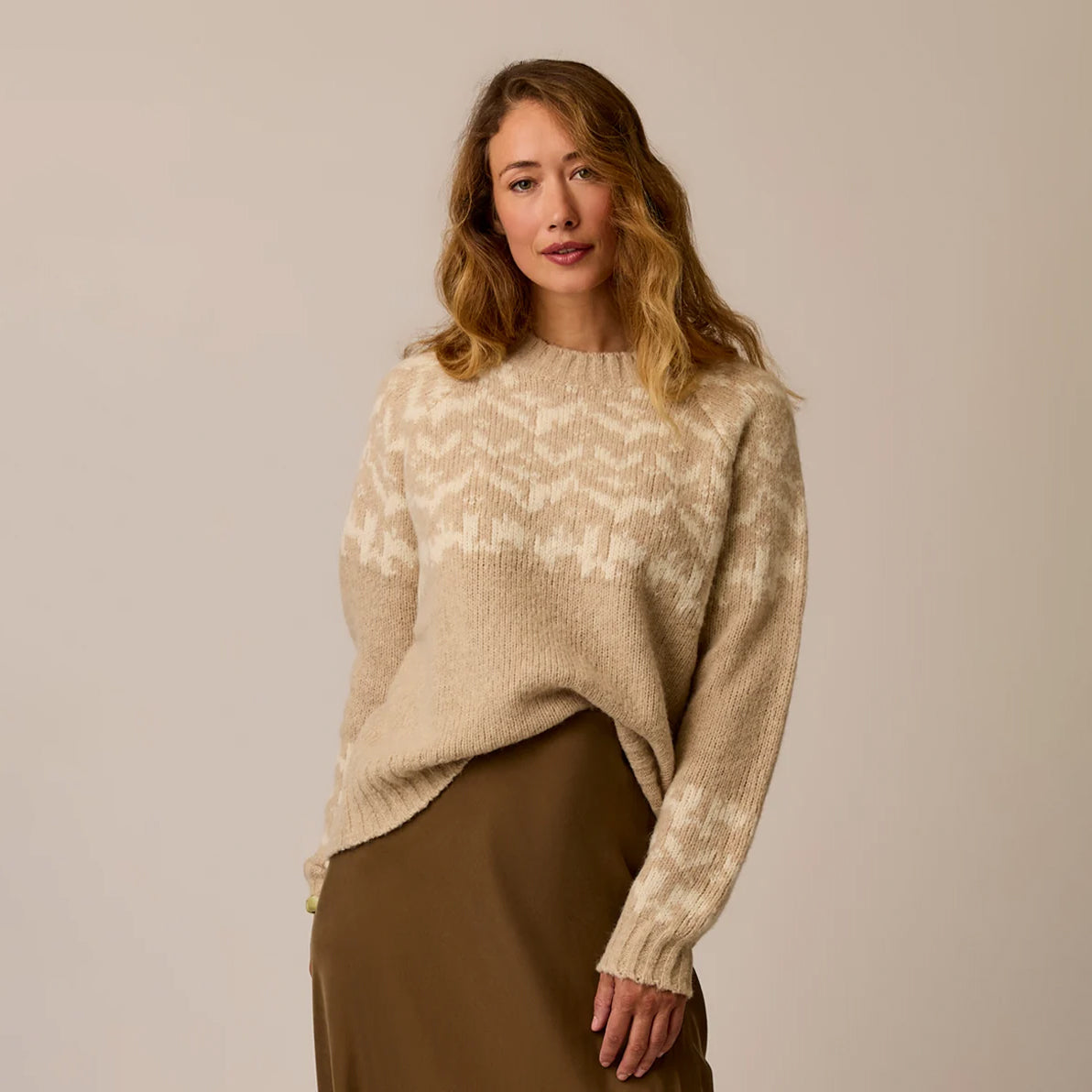 A woman wearing a beige and cream patterned knit sweater with a brown skirt, standing against a plain beige studio background.