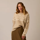 A woman wearing a beige and cream patterned knit sweater with a brown skirt, standing against a plain beige studio background.