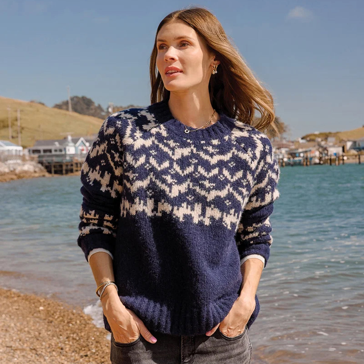 A woman wearing a navy and cream patterned knit sweater stands on a beach with the ocean and hills in the background.