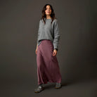 A woman stands against a dark gray studio background wearing a gray knit sweater, a long dusty mauve wrap-style maxi skirt, and gray patterned sandals.