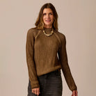 A woman modeling a brown ribbed knit mock neck sweater with raglan sleeves, paired with dark gray jeans and a chunky beaded necklace, against a solid beige background.