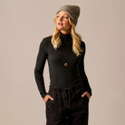 A woman wearing a black long-sleeve mock neck top, black drawstring sweatpants, a gray knit beanie, and a black pendant necklace, posing against a solid beige background.
