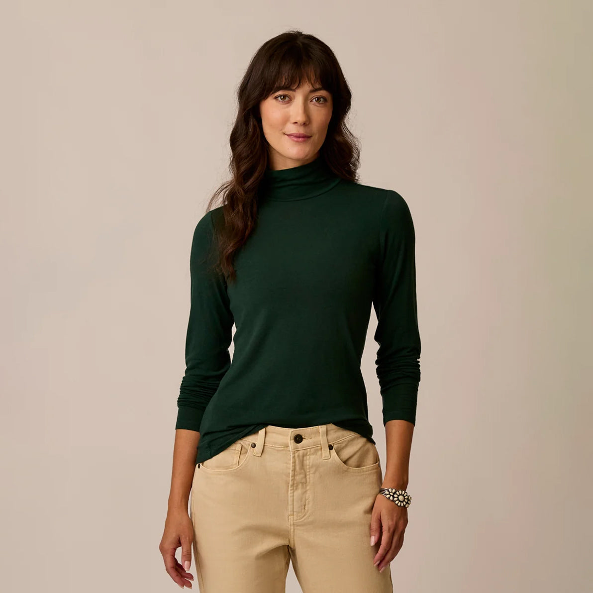 A woman modeling a dark forest green long-sleeve mock neck turtleneck sweater, paired with beige pants, against a solid light beige background.