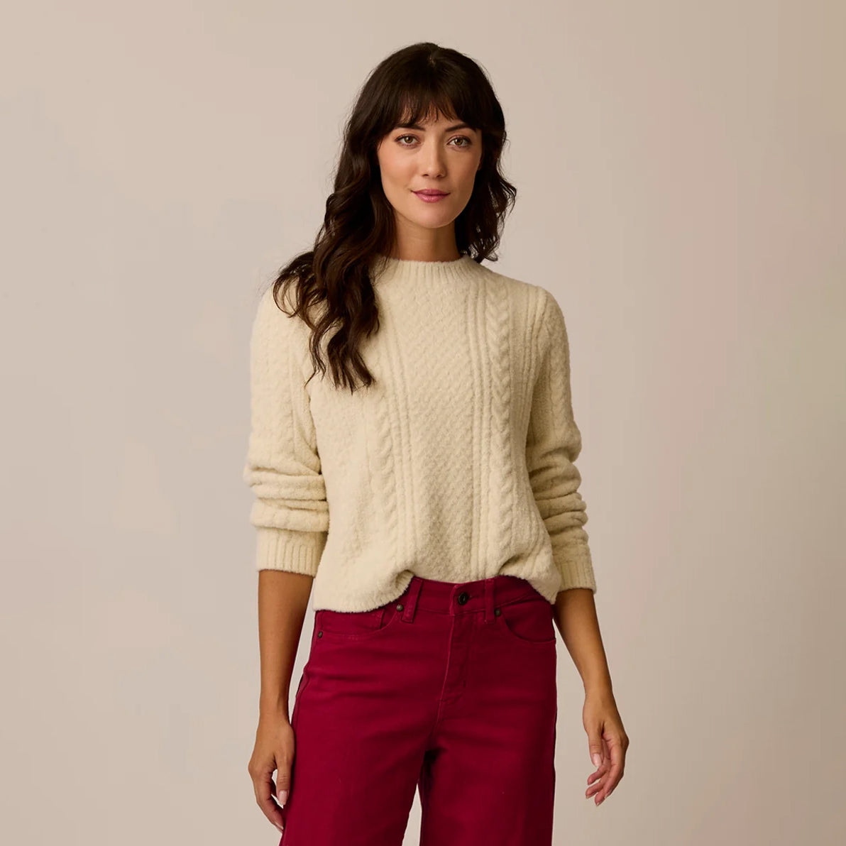 A woman modeling a cream cable-knit sweater with three-quarter sleeves, paired with dark red pants, against a solid light beige background.