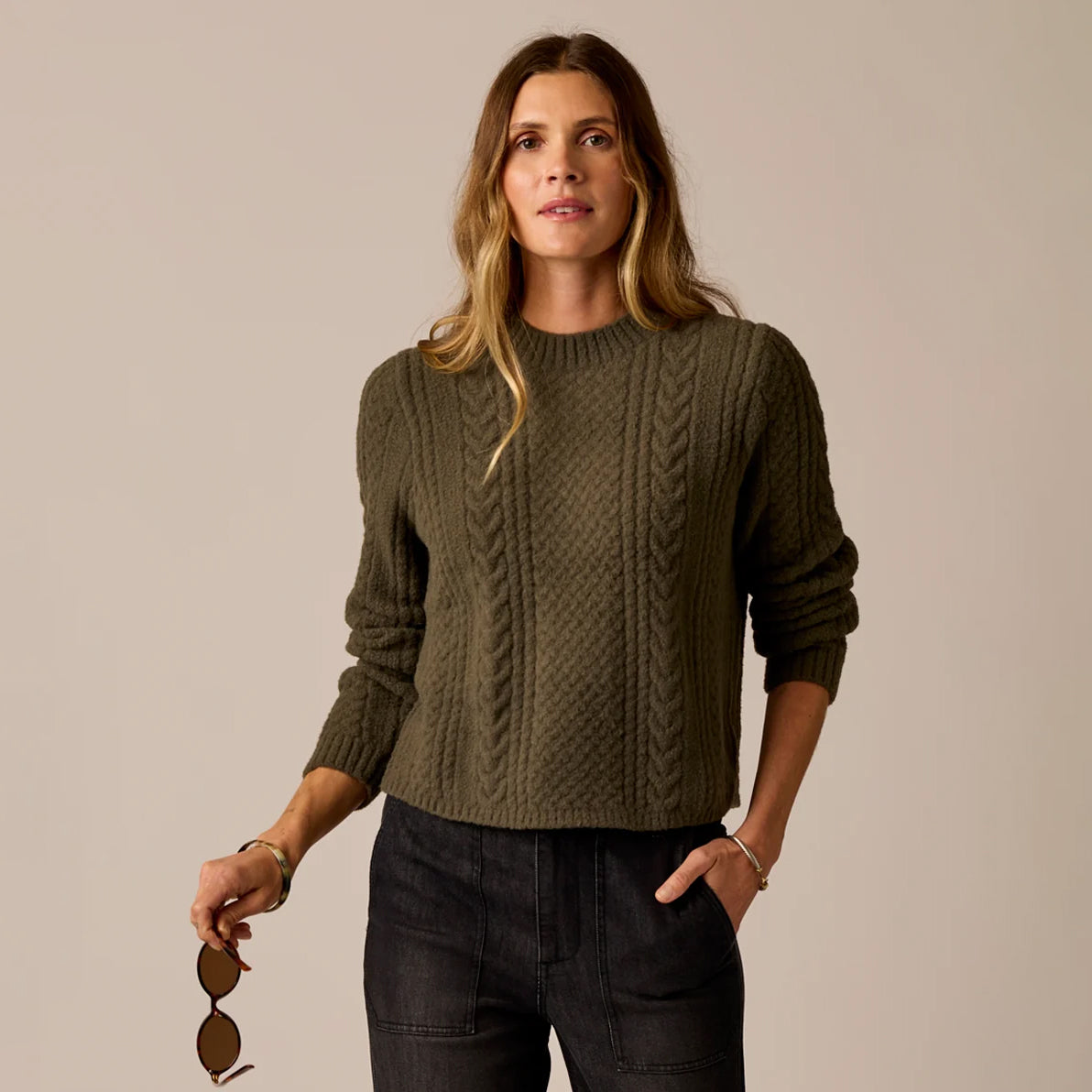 A woman wearing an olive green cable-knit crewneck sweater and dark jeans, holding a pair of sunglasses against a plain beige studio background.