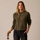 A woman wearing an olive green cable-knit crewneck sweater and dark jeans, holding a pair of sunglasses against a plain beige studio background.