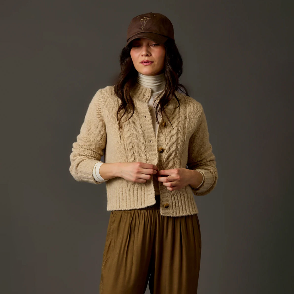 A woman wearing a beige cable-knit cardigan over a white turtleneck, paired with brown wide-leg pants and a brown cap, standing against a dark gray studio background.