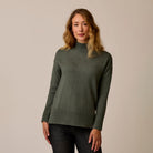 A woman wearing a muted olive green mock neck knit sweater with ribbed hem and cuffs, paired with dark jeans, standing against a plain beige studio background.