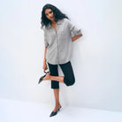 A woman posing in a light gray button-down shirt, black cropped capri pants, and black pointed-toe mule heels against a clean white studio background.