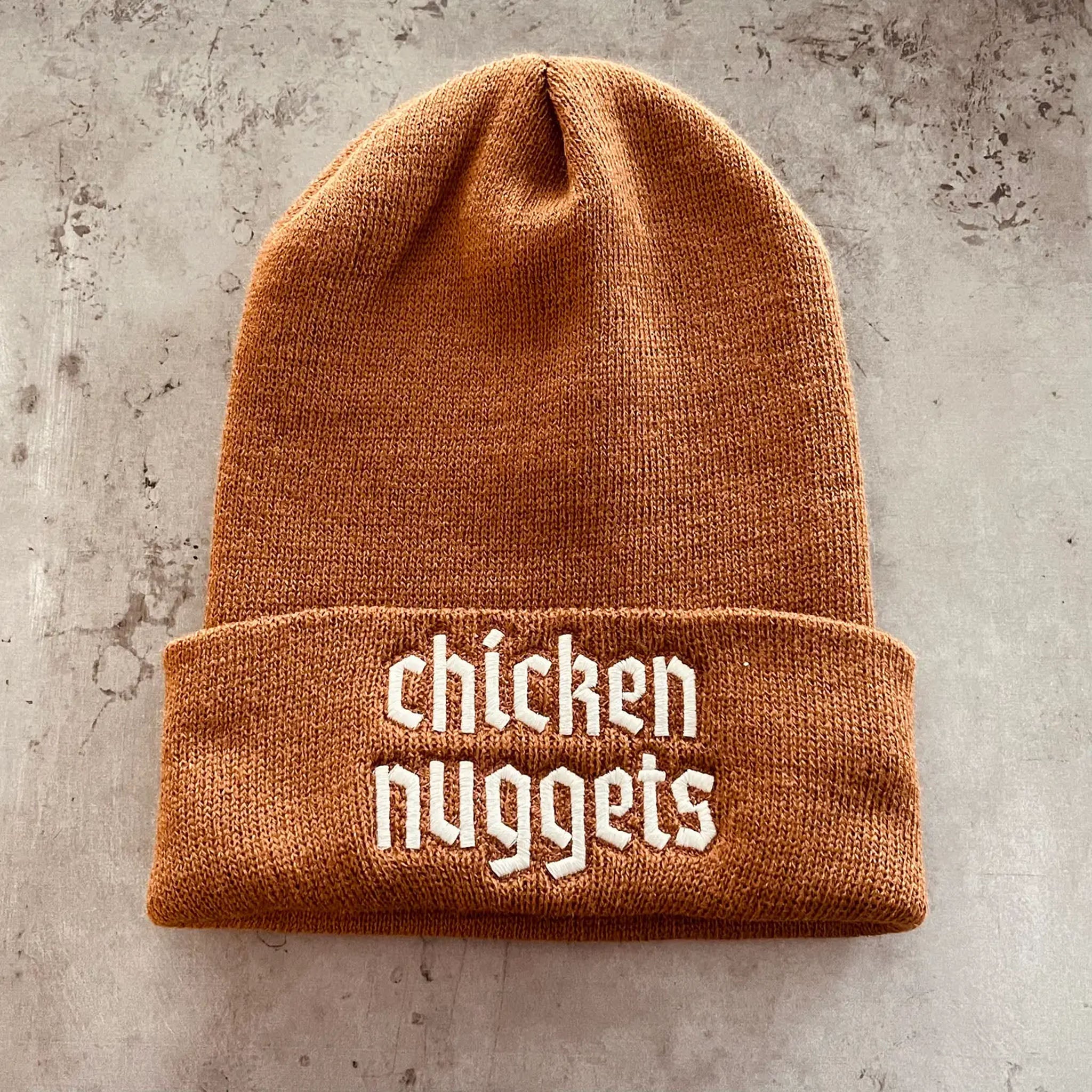 Brown beanie with 'chicken nuggets' text on a concrete surface