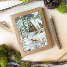 Winter-themed greeting card with a snowman, fox, and cabin design on a natural background.