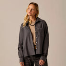 A woman modeling a dark charcoal gray button-up shirt with a chest pocket, worn open over a beige knit top and dark jeans, set against a solid light beige studio background.