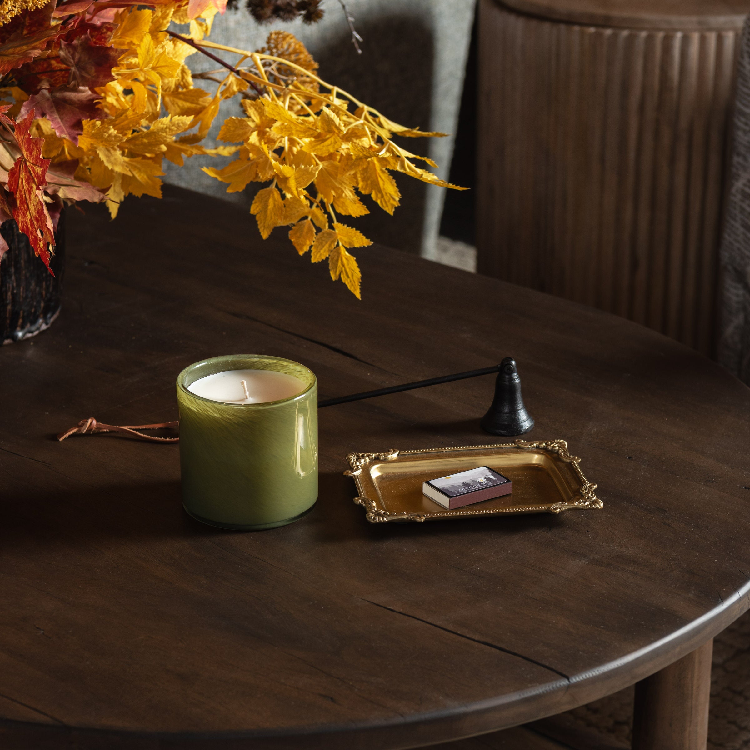 A green glass candle jar with white wax sits on a dark wooden table next to a small gold tray holding a matchbox, with a black candle snuffer and autumn leaves in the background.