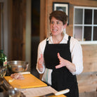 A woman with short brown hair wearing a white button-down shirt and a black apron stands in a rustic kitchen, gesturing with her hands while speaking. In the foreground, yellow cutting boards, a metal bowl, and chopped green herbs are visible on a counter.