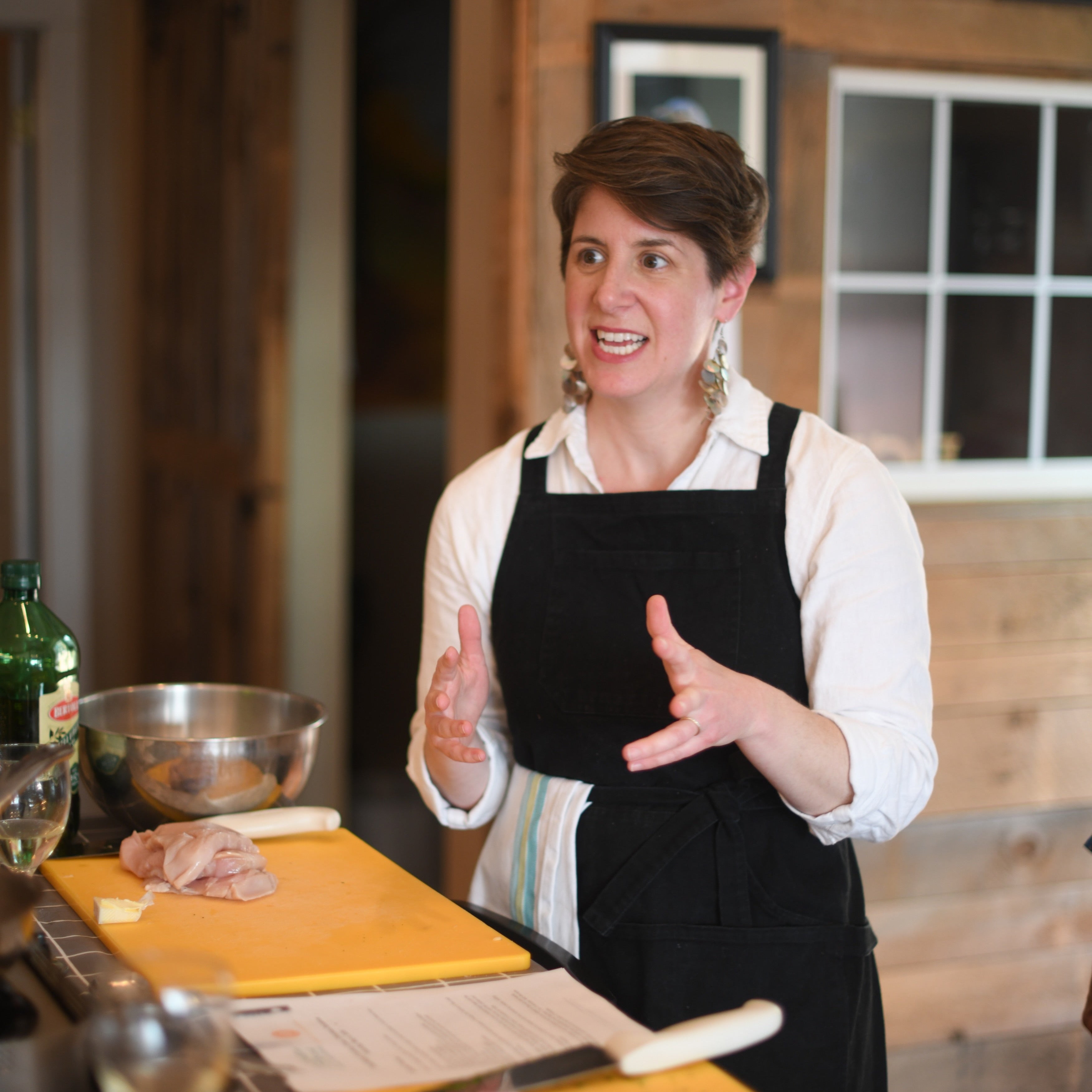 A woman with short brown hair wearing a white button-down shirt and a black apron stands in a rustic kitchen, gesturing with her hands while speaking. In the foreground, yellow cutting boards, a metal bowl, and chopped green herbs are visible on a counter.