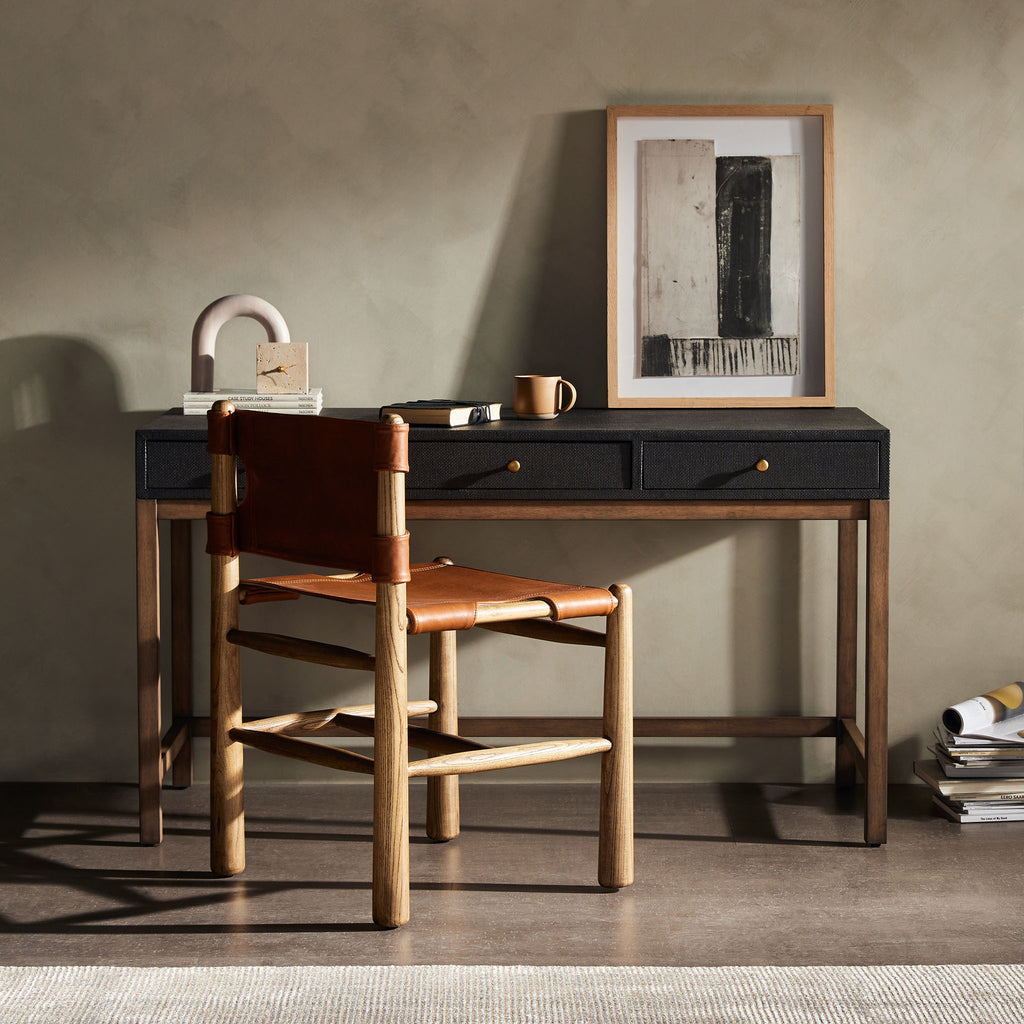 Four Hands Fiona Desk in Black Raffia with brass legs in a room with grey walls and a leather chair 