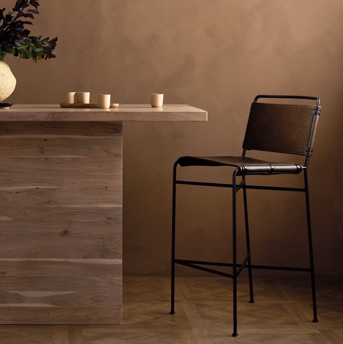 Four Hands Wharton Bar Stool in Distressed Brown next to a wood table at  Addison West