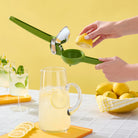 Person using a green manual lemon squeezer with a glass of lemonade and lemons on a yellow background.