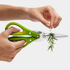 Green kitchen shears being used to trim a sprig of rosemary on a white background