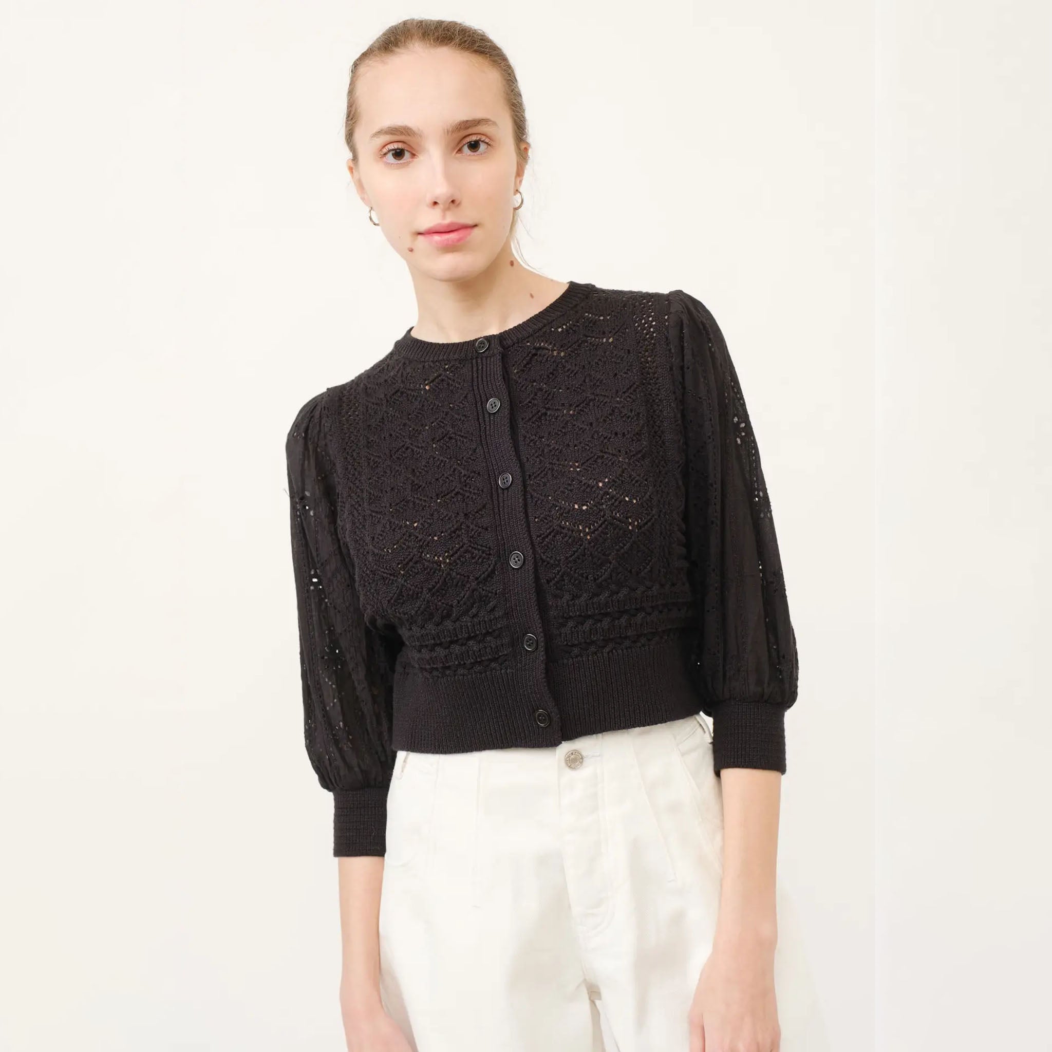 Woman wearing an aaron and amber black knit cardigan over a white outfit on a plain background