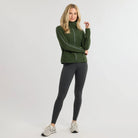 Woman wearing green sweater with dark colored pants on a plain background.