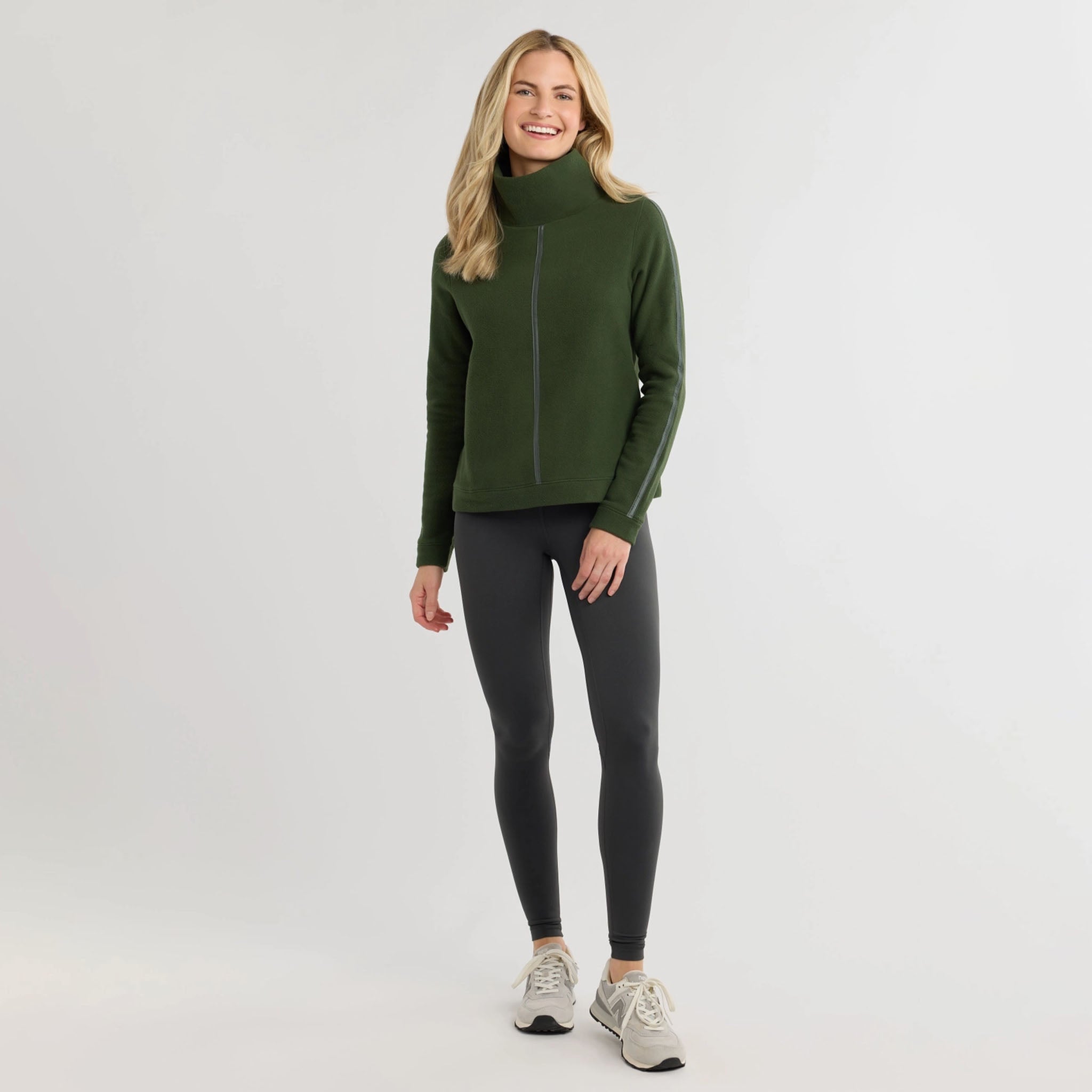 Woman wearing green sweater with dark colored pants on a plain background.