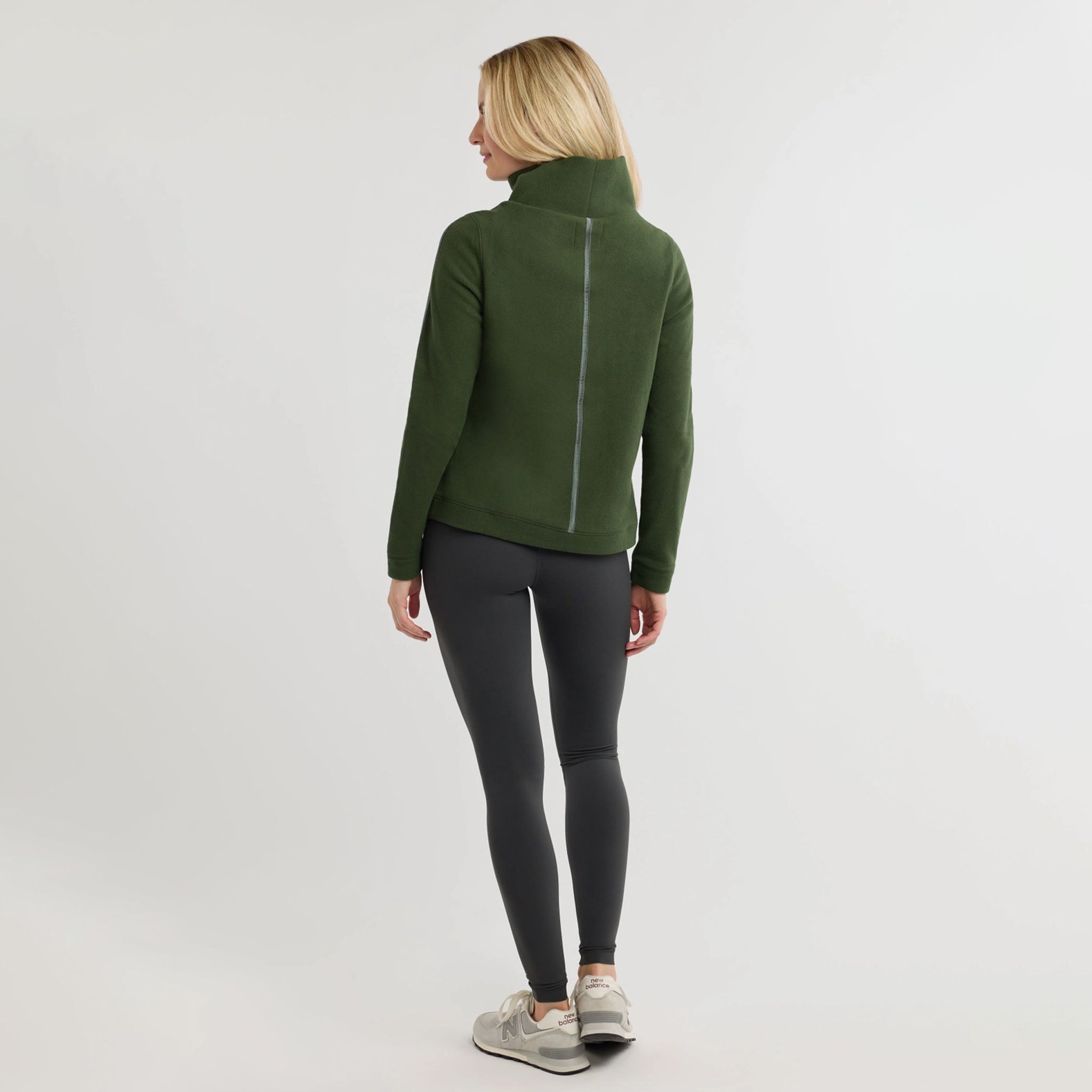 Woman wearing green sweater with dark colored pants on a plain background.