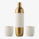 A stainless steel flask with gold and white coloring, accompanied by two white tumblers, against a white background.