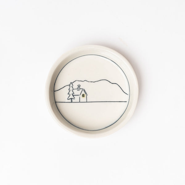 Small white handmade dish with drawing of home and mountains on a white background