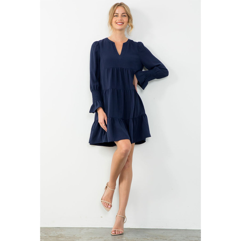 Blond model wearing THML Long Sleeve Tiered Midi Dress in Navy standing in front of a white wall