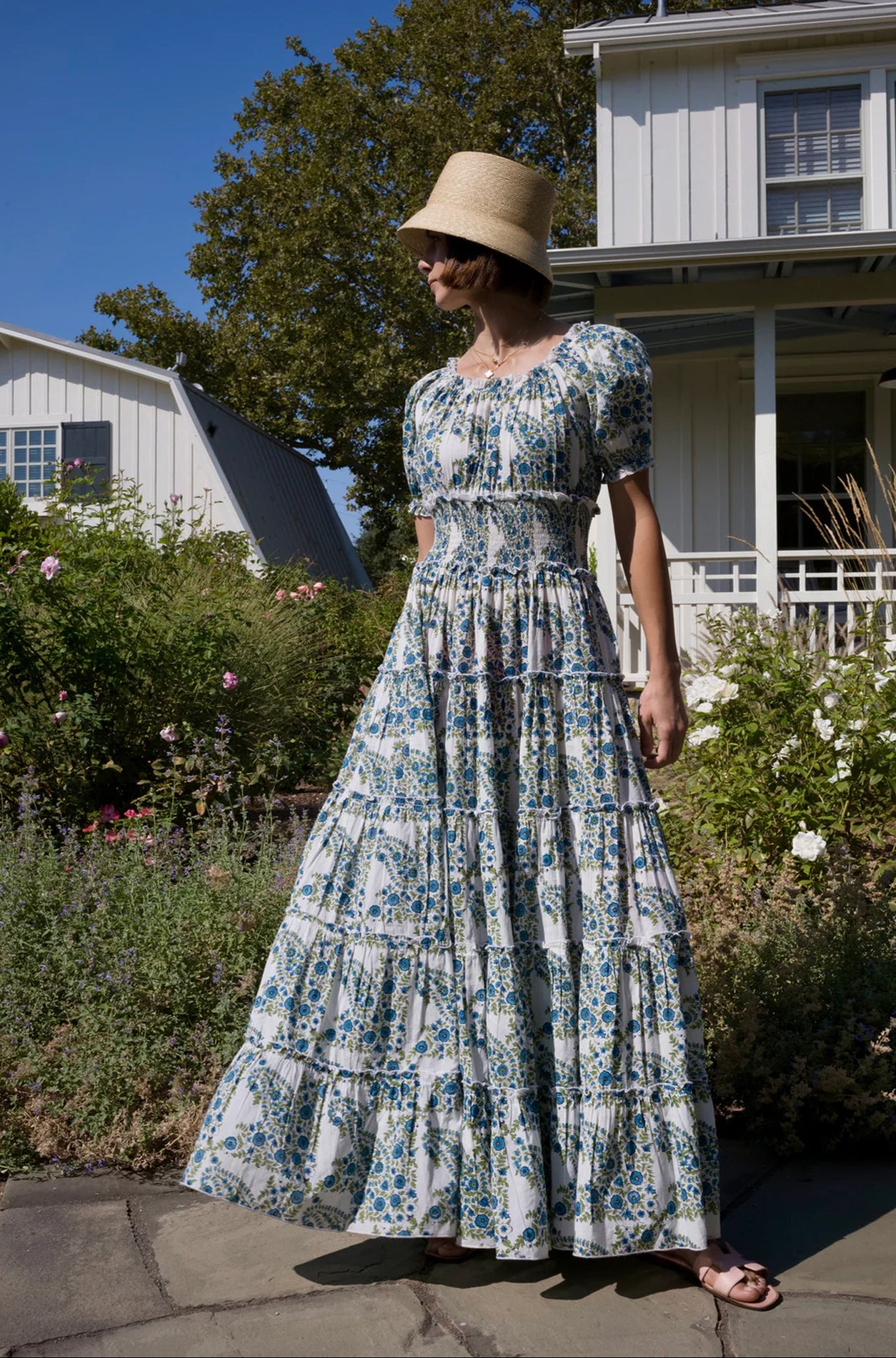 Model wearing Maison Marigold Santorini Maxi Dress in Blue/Green in front of a white house with flowers
