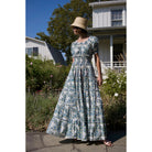Model wearing Maison Marigold Santorini Maxi Dress in Blue/Green in front of a white house with flowers