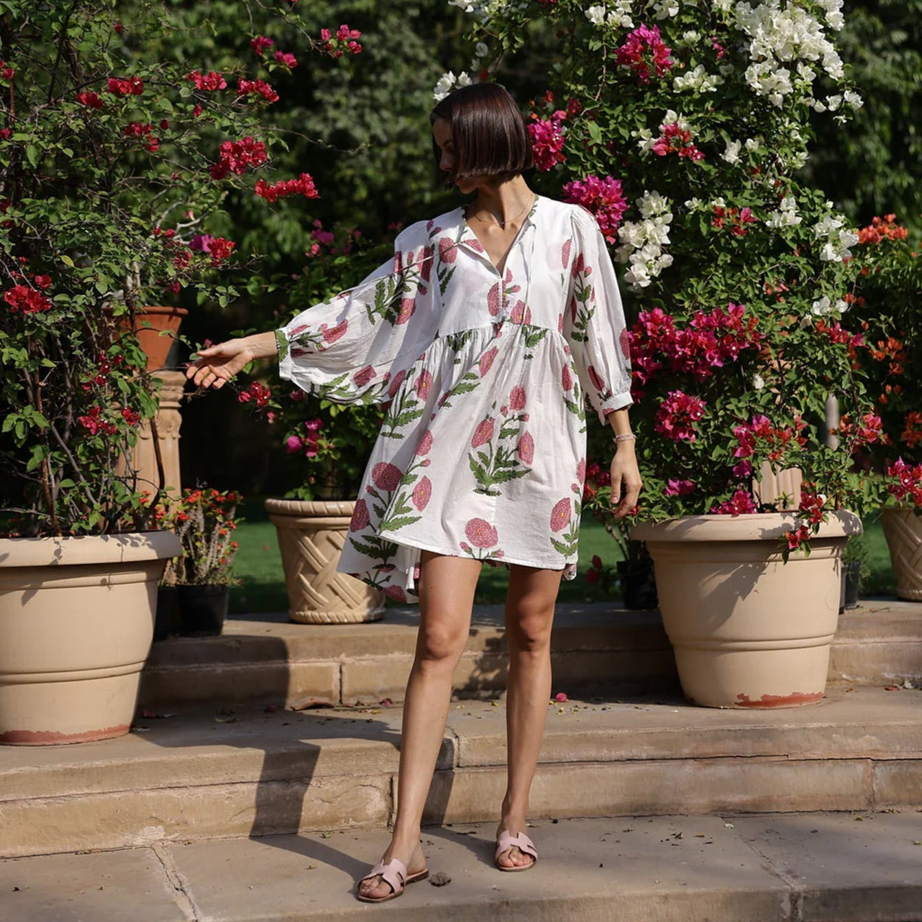 Model wearing Maison Marigold Taj Mini Dress in Pink on stone steps with flowering plants in the background