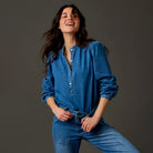 A woman wearing a blue denim long-sleeve button-up shirt and matching blue denim jeans, posing against a dark gray studio background.