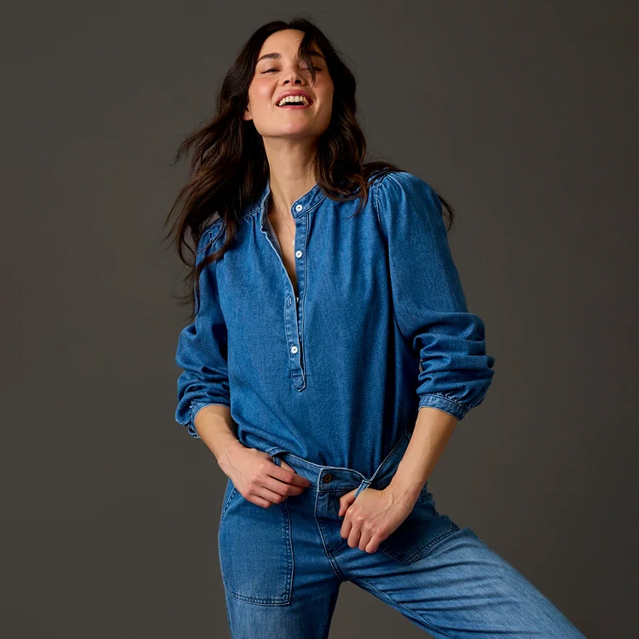 A woman wearing a blue denim long-sleeve button-up shirt and matching blue denim jeans, posing against a dark gray studio background.