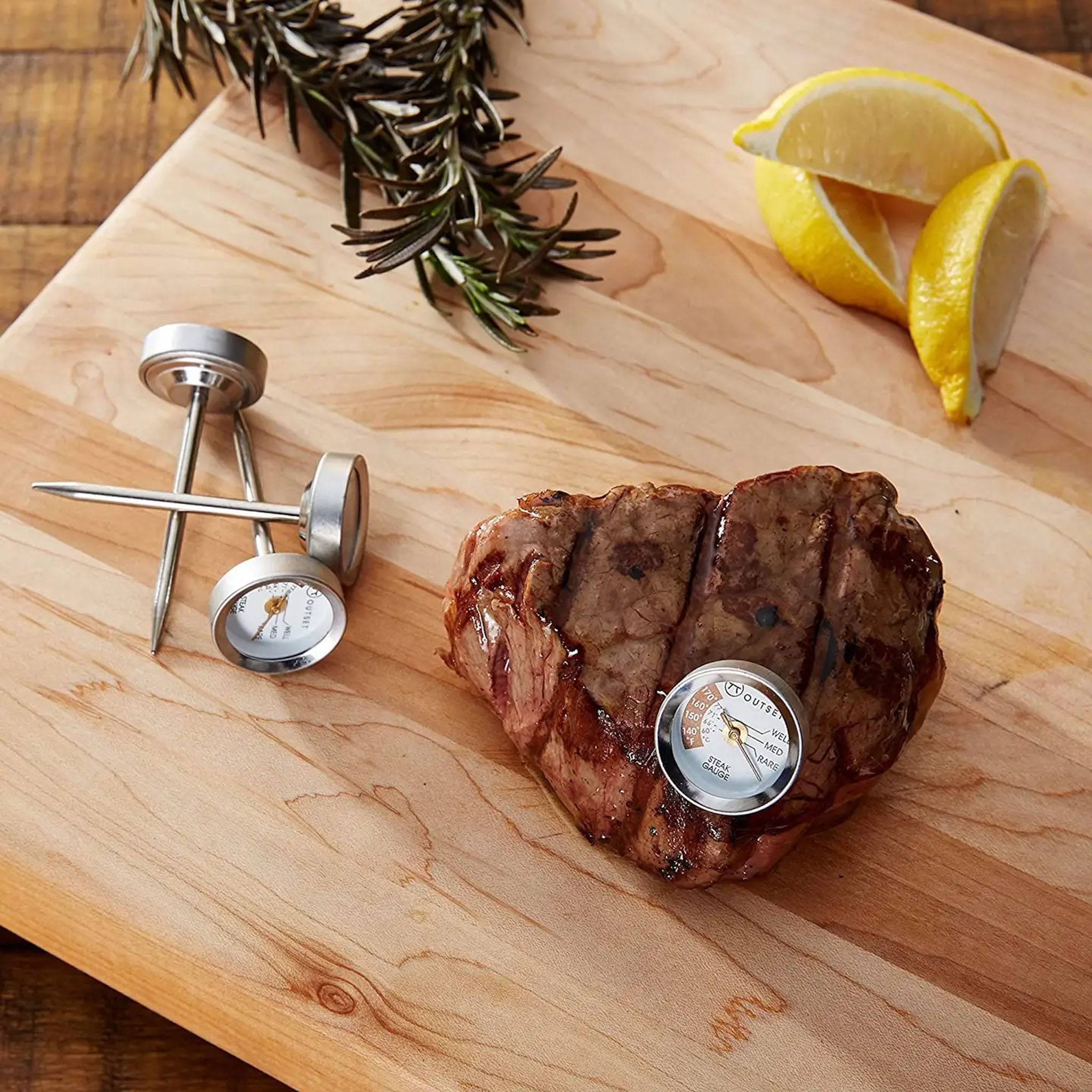Steak with a thermometer on a wooden cutting board, surrounded by rosemary and lemon slices.