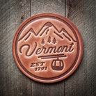 A brown leather coaster with embossed text and a ski gondola design, on a wooden background.