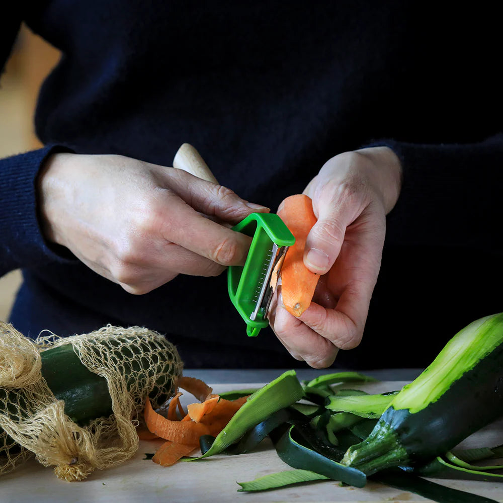 Person using a green vegetable peeler on carrots with zucchini and a mesh bag in the foreground.