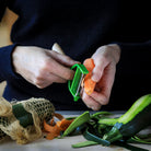 Person using a green vegetable peeler on carrots with zucchini and a mesh bag in the foreground.