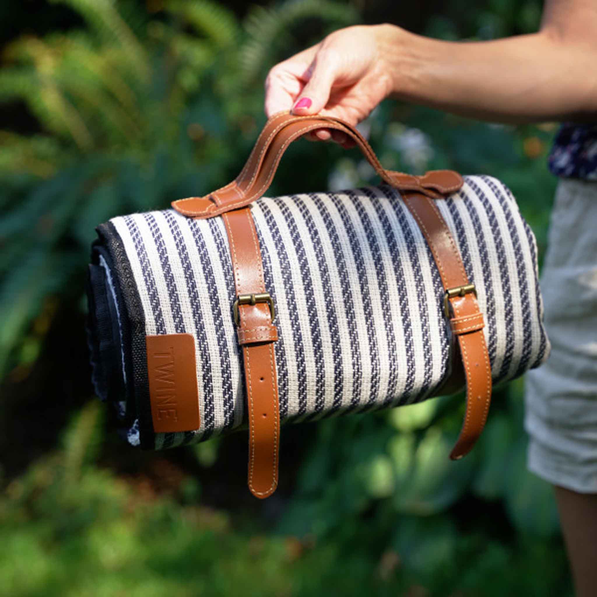 Picnic blanket rolled up and shown with leather handle held by a woman in a wooded setting 
