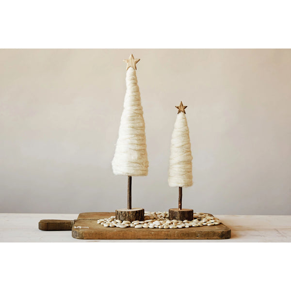 Two white wool christmas trees with gold stars and wood bases on a wood tray in front of a white wall