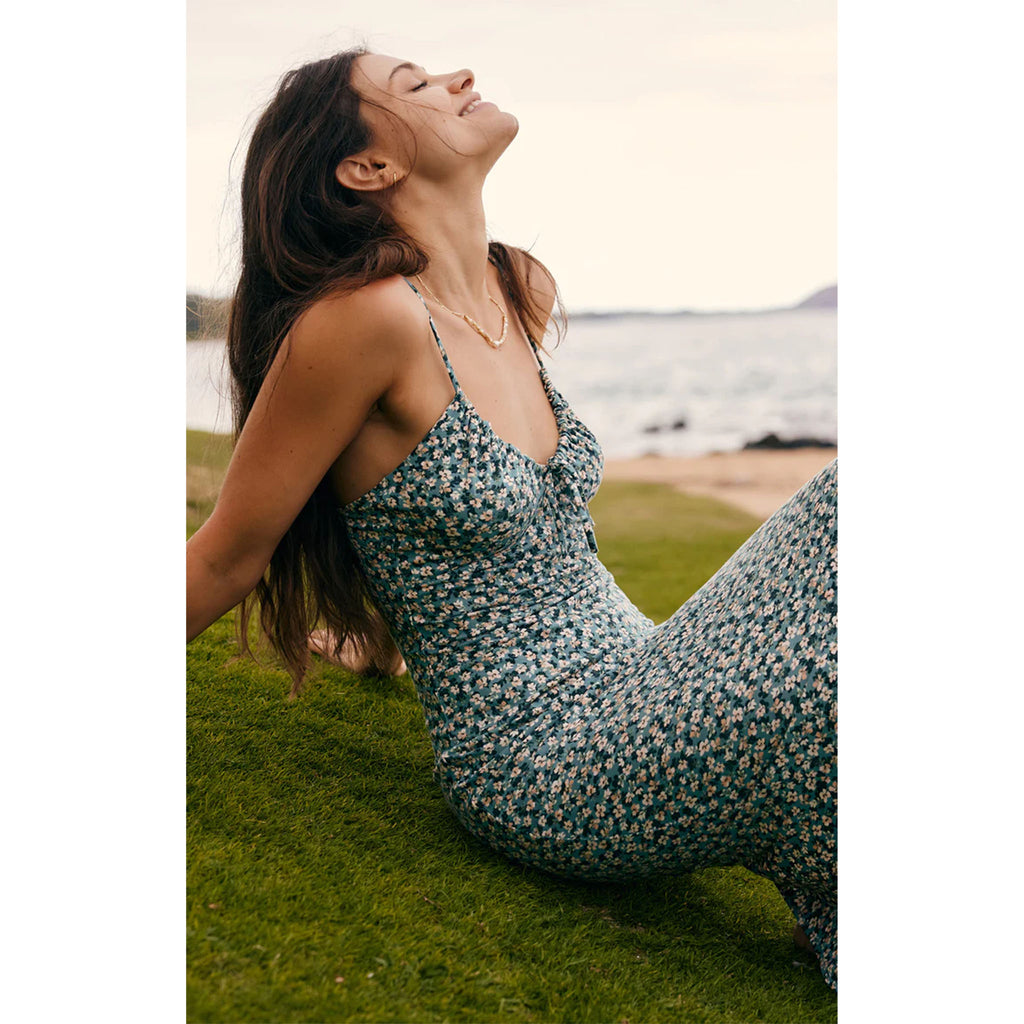 Model wearing Z Supply Melinda Ditsy Floral Midi Dress In Emerald Isle  sitting on grass by on a beach with ocean in the background