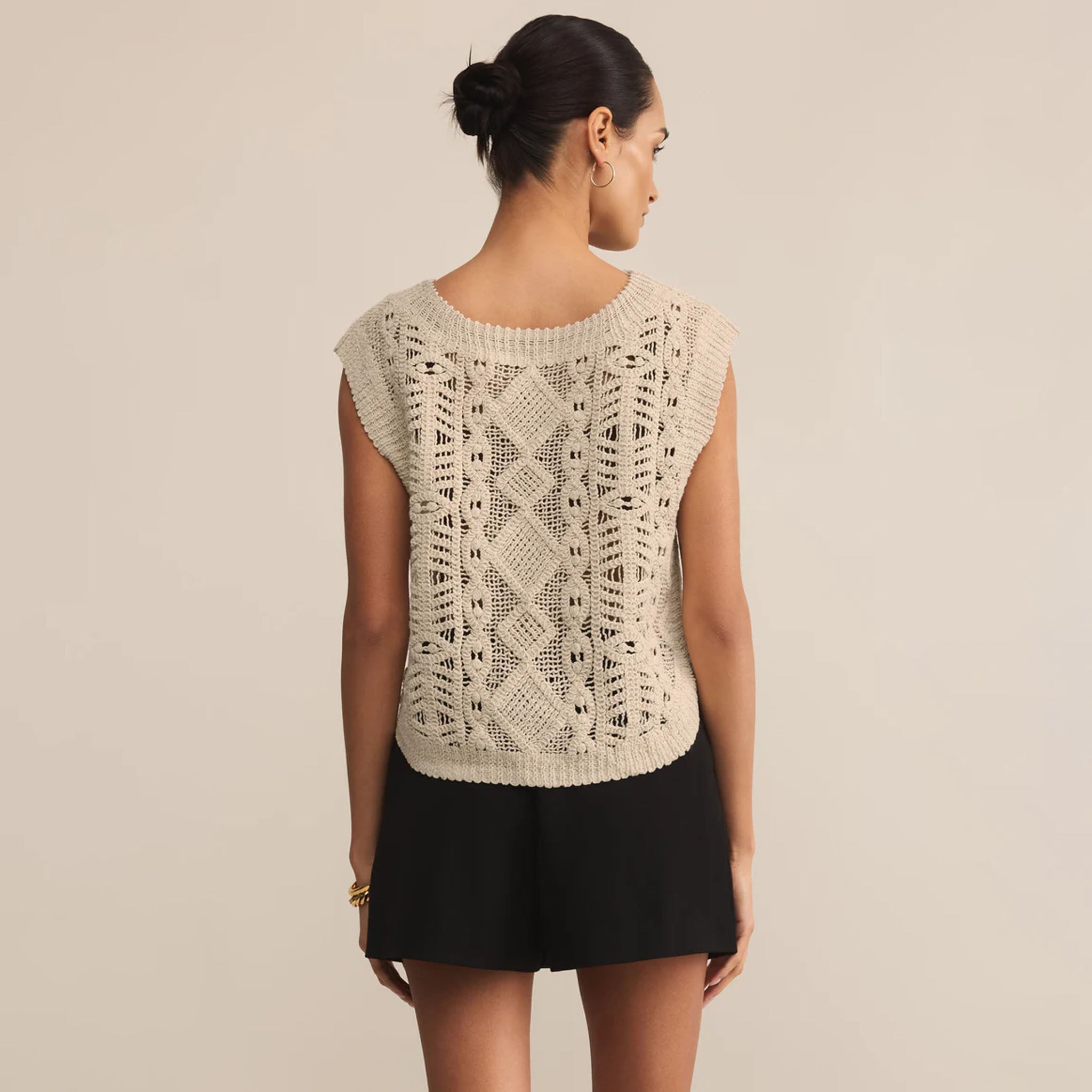 Woman wearing a beige sleeveless knitted top with a textured pattern on a plain background