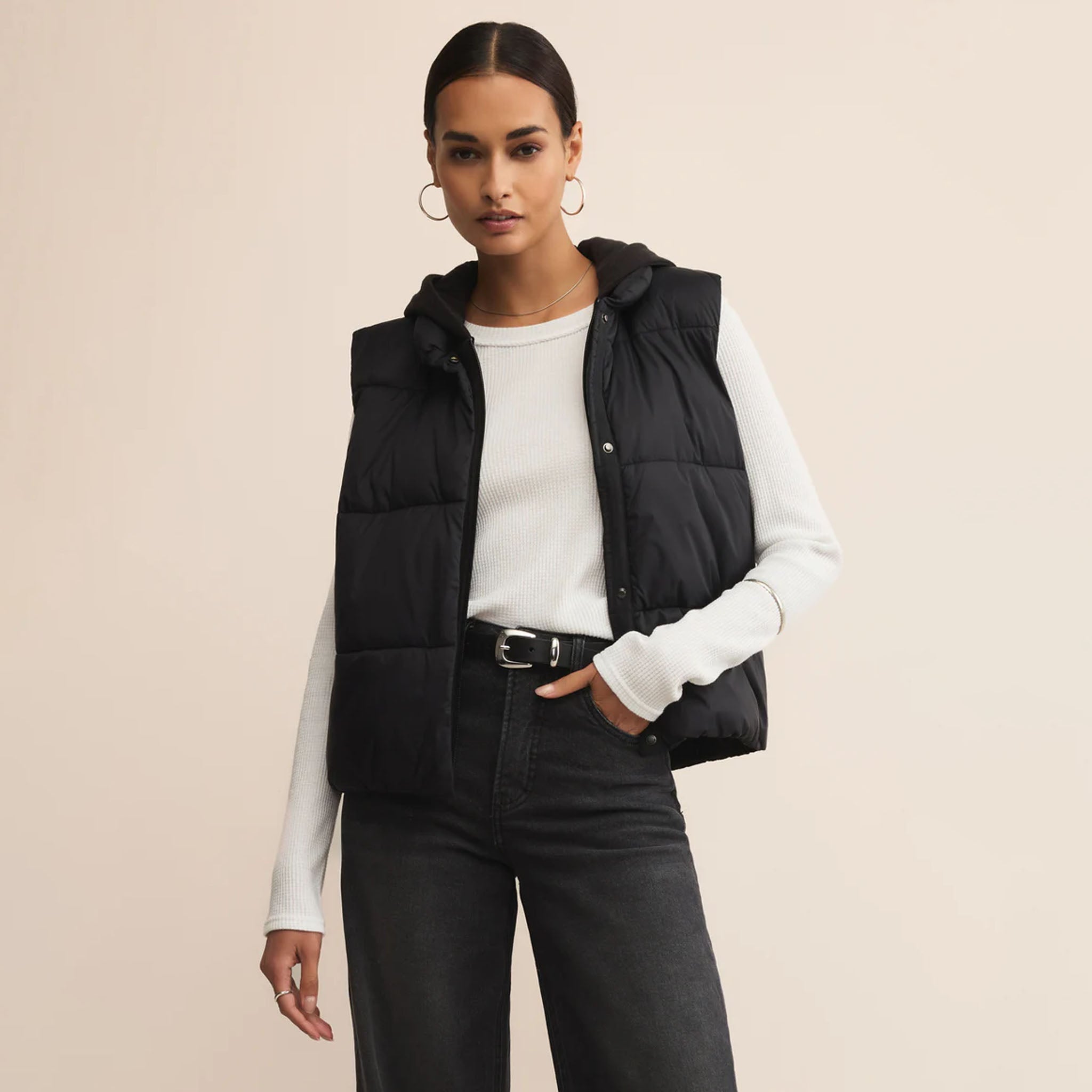 Woman wearing a black puffer vest over a white long-sleeve shirt on a beige background