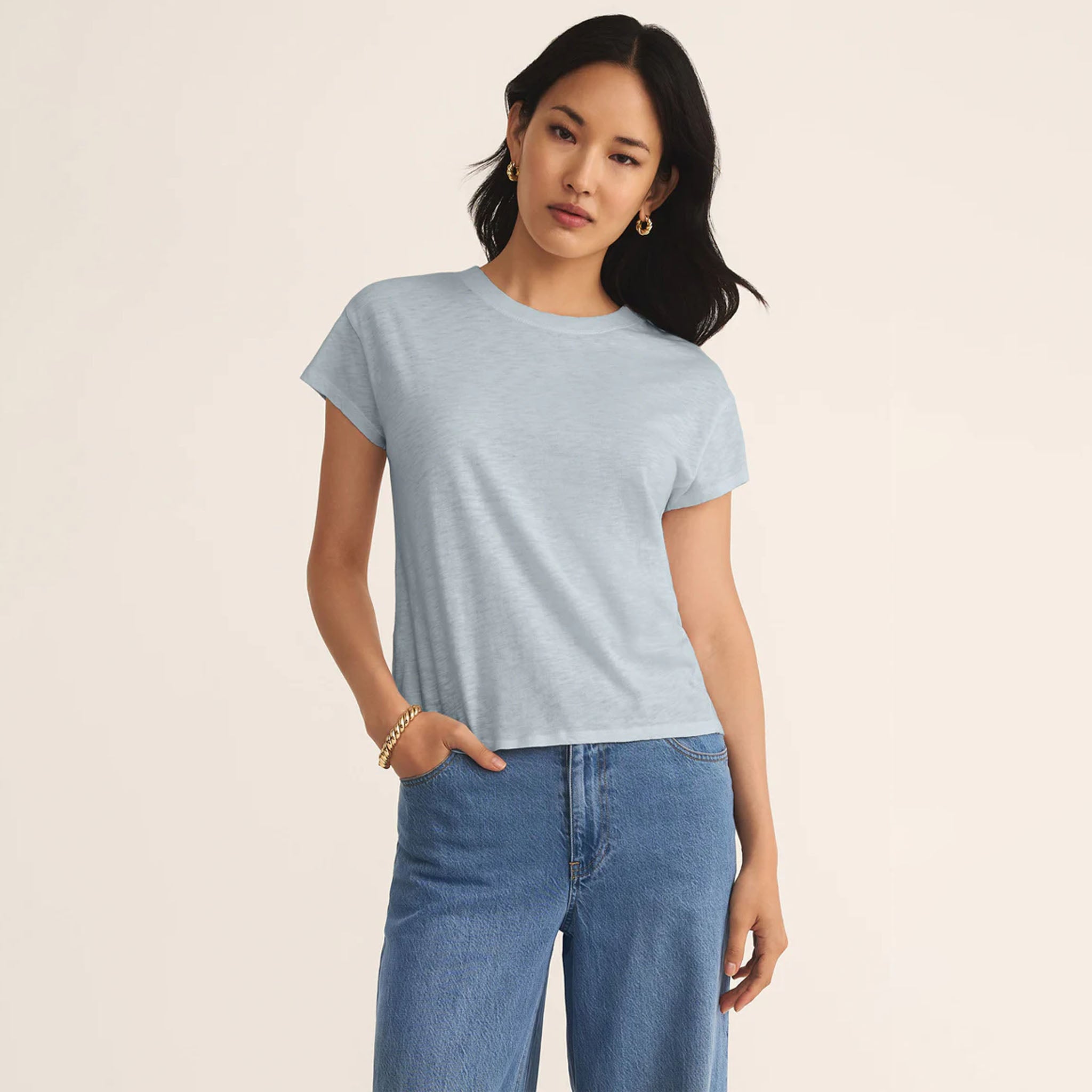 Woman wearing a light blue t-shirt, the Z Supply Modern Slub Tee in Clearwater, and jeans on a beige background